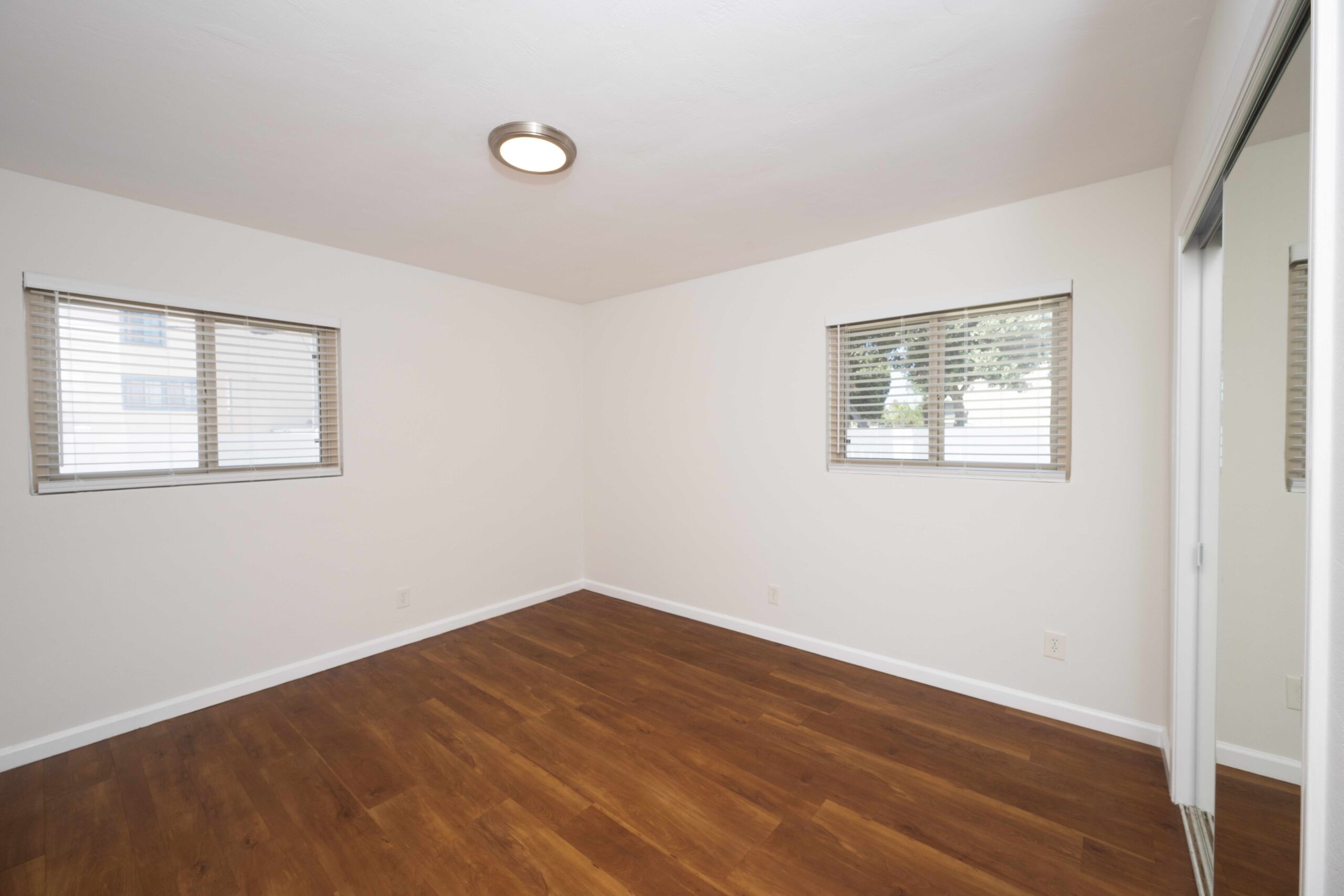 Gallery Bright bedroom with wood-look flooring, two windows, and mirrored wardrobe doors.