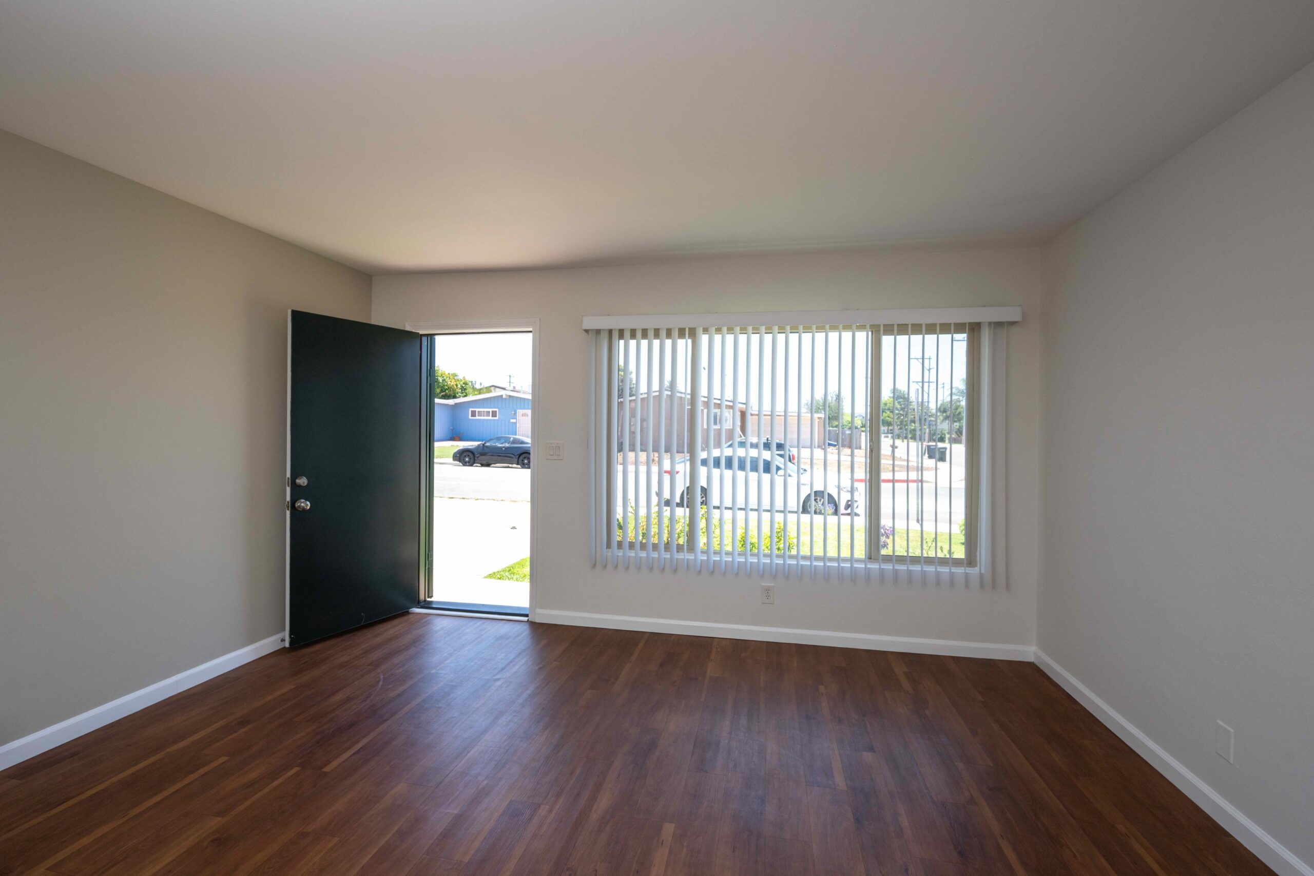 Gallery Renovated living room with wood-look flooring, front entry, and vertical blinds.